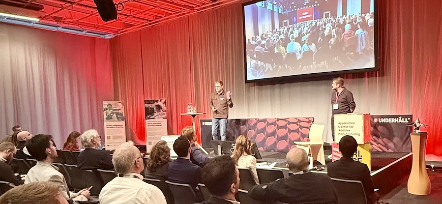 Två personer håller föredrag på en scen med publik framför sig.