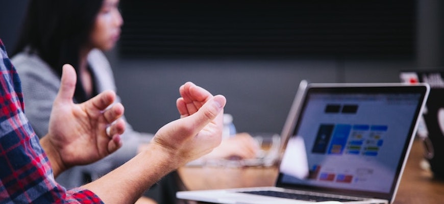Två händer som gestikulerar till vänster i bild framför en bärbar dator med ett digitalt möte igång. En kvinna i bakgrunden.
