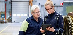 Två kvinnor står och tittar ner i en iPad ute i en industrilokal.